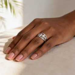 Bague Guadeloupe 7 Main de femme portant la bague Guadeloupe couleur argent, un bijou en acier inoxydable de 13 mm de large, avec un motif ajouré représentant la carte des Antilles, des palmiers et une silhouette tropicale, posée sur un tissu beige dans une lumière naturelle.