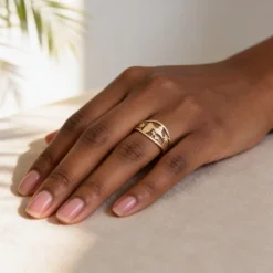 Bague Guadeloupe 6 Main de femme portant la bague Guadeloupe dorée, un bijou en acier inoxydable doré de 13 mm de large, avec un motif ajouré représentant la carte des Antilles, des palmiers et une silhouette tropicale, posée sur un tissu beige dans une lumière naturelle.
