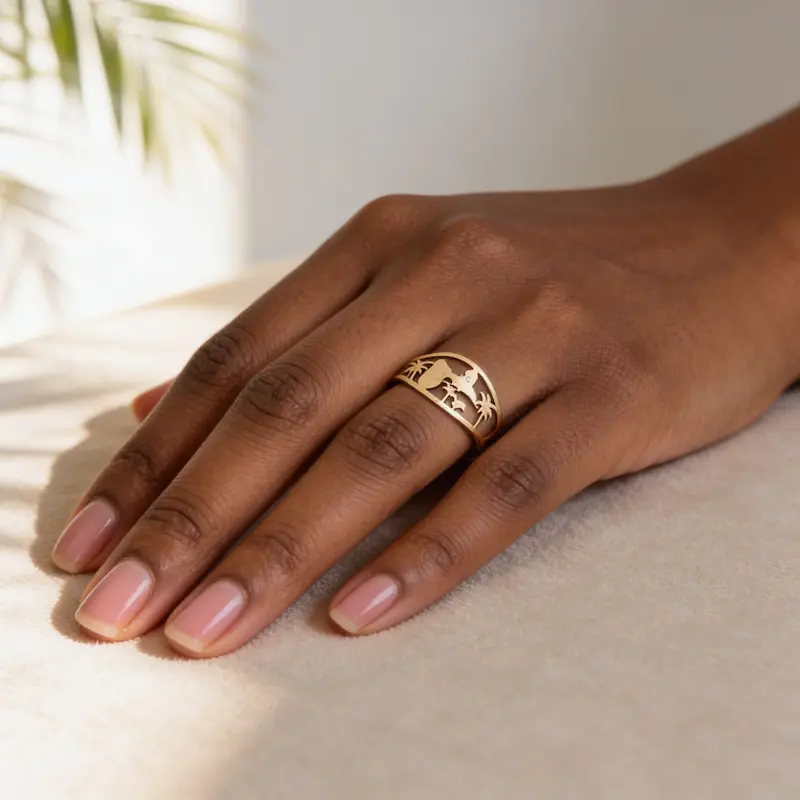 Bague Guadeloupe 3 Main de femme portant la bague Guadeloupe dorée, un bijou en acier inoxydable doré de 13 mm de large, avec un motif ajouré représentant la carte des Antilles, des palmiers et une silhouette tropicale, posée sur un tissu beige dans une lumière naturelle.