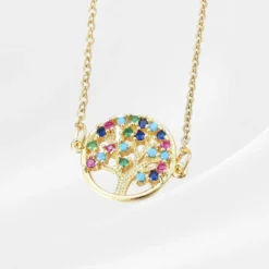 Pendentif Arbre de vie 13 Collier Arbre de Vie en acier inoxydable plaqué or, médaillon rond ajouré fixé latéralement sur une chaîne forçat dorée traversante, avec un arbre aux branches texturées orné de grands zircons ronds multicolores (bleu saphir, turquoise, rose fuchsia, vert émeraude, orange) répartis en couronne dense sur les branches, photographié à plat sur fond blanc satiné.