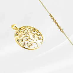 Pendentif Arbre de vie 11 Pendentif Arbre de Vie en acier inoxydable plaqué or, grand médaillon rond ajouré à bordure pleine avec un arbre aux branches fines et ramifiées ornées de petites fleurs serties de zircons multicolores épars (rouge, vert, bleu, mauve, blanc), photographié en biais prononcé sur fond gris clair satiné, avec une fine chaîne dorée forçat visible en arrière-plan.