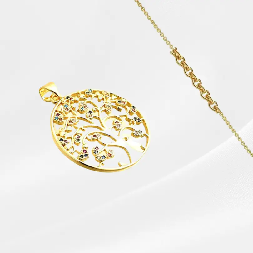 Pendentif Arbre de vie 5 Pendentif Arbre de Vie en acier inoxydable plaqué or, grand médaillon rond ajouré à bordure pleine avec un arbre aux branches fines et ramifiées ornées de petites fleurs serties de zircons multicolores épars (rouge, vert, bleu, mauve, blanc), photographié en biais prononcé sur fond gris clair satiné, avec une fine chaîne dorée forçat visible en arrière-plan.