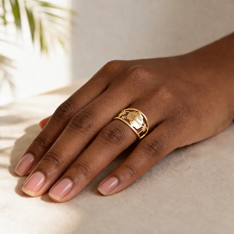 Bague La Réunion 3 Main de femme à la peau foncée portant la bague La Réunion en acier inoxydable doré, un bijou de 13 mm de large avec un motif ajouré représentant la carte de l'île de La Réunion, des palmiers et les inscriptions "LA RÉUNION" et "CILAOS", posée sur un tissu beige dans une lumière naturelle douce avec ombre de feuille de palmier en arrière-plan.