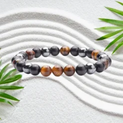 Bracelet hématite en pierres naturelles noires et œil de tigre brun doré, perles rondes de 8 mm montées sur élastique, posé sur sable blanc zen avec feuilles de bambou — bijou unisexe homme femme