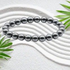 Bracelet en pierres naturelles d'hématite gris argenté brillant, perles rondes de 8 mm montées sur élastique, posé sur sable blanc zen avec feuilles de bambou — bijou unisexe homme femme