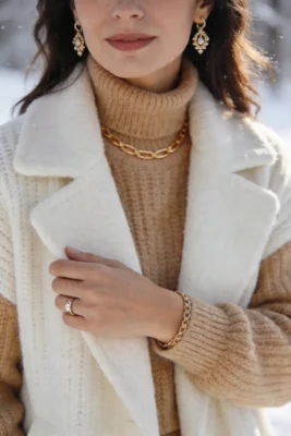 Quels bijoux porter selon les saisons ? 4 Femme portant des bijoux dorés avec tenue d’hiver en maille beige, collier visible, boucles d’oreilles fines et ambiance lumineuse de saison