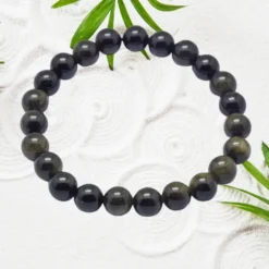 Bracelet en Obsidienne noire naturelle, perles rondes polies 10 mm, monté sur élastique, posé sur fond blanc texturé avec feuilles vertes — bijou pierre volcanique homme femme