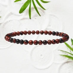 Bracelet en Obsidienne 5 Bracelet obsidienne acajou femme, petites perles rondes rouge brun et noir 4 mm sur élastique fin, fond sable blanc texturé avec feuilles vertes — bijou pierre naturelle fin poignet
