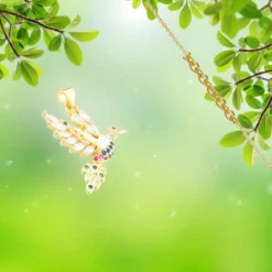Pendentif Oiseau en métal doré à l'or fin, serti de zirconiums multicolores (blanc, bleu, rose, vert), représentant un colibri aux ailes déployées — bijou femme délicat sur fond naturel vert