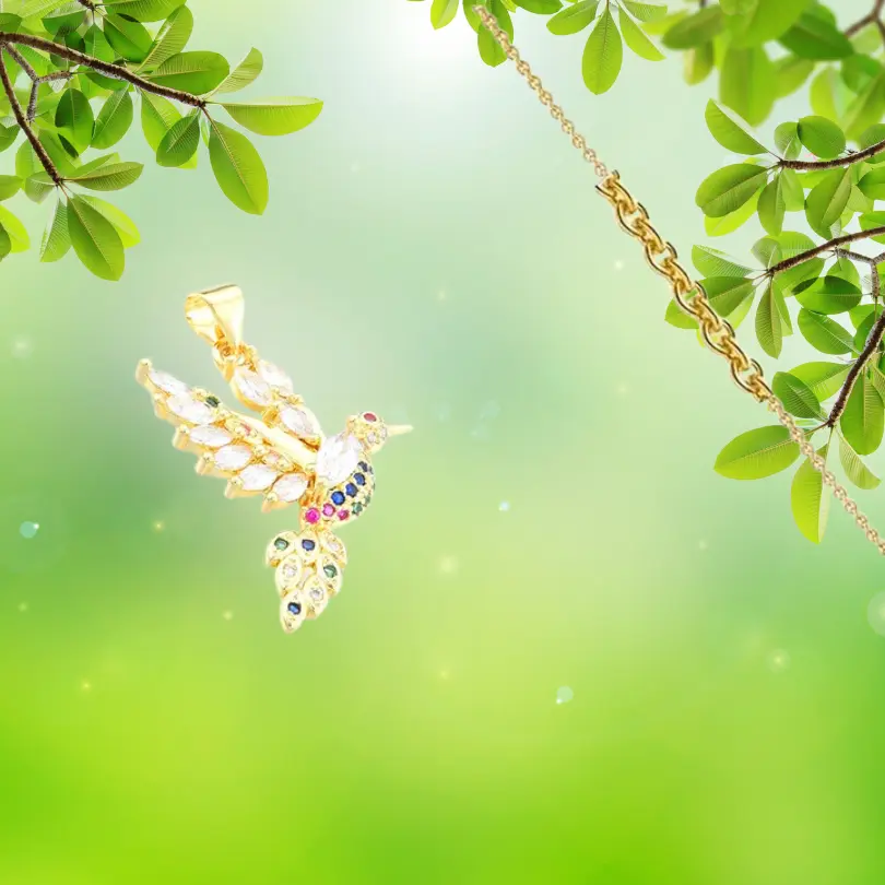 Pendentif Oiseau 1 Pendentif Oiseau en métal doré à l'or fin, serti de zirconiums multicolores (blanc, bleu, rose, vert), représentant un colibri aux ailes déployées — bijou femme délicat sur fond naturel vert
