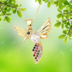 Pendentif oiseau doré à l'or fin avec zirconiums multicolores (rose, bleu, vert, violet, noir) pavés sur le corps du colibri — collier bijou femme suspendu sur fond naturel vert, chaîne dorée visible