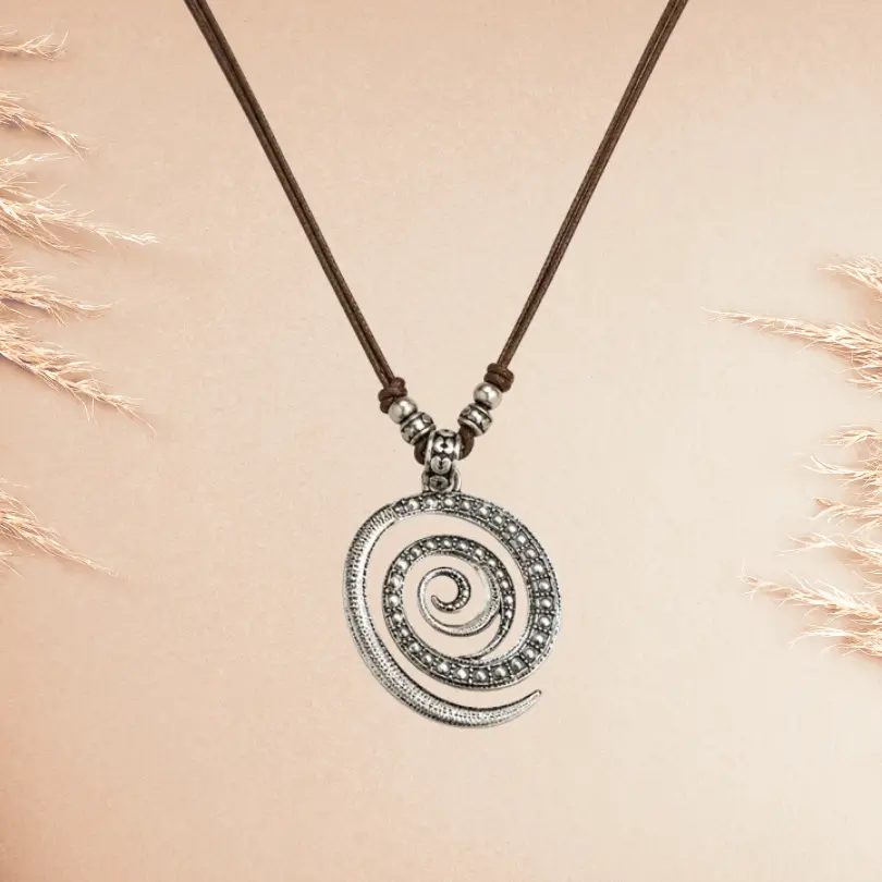 Collier Bohème 1 Collier Bohème pendentif spirale en métal argenté vieilli sur cordon cuir marron ajustable, photographié sur fond beige rosé avec épis de pampa — bijou fantaisie ethnique femme