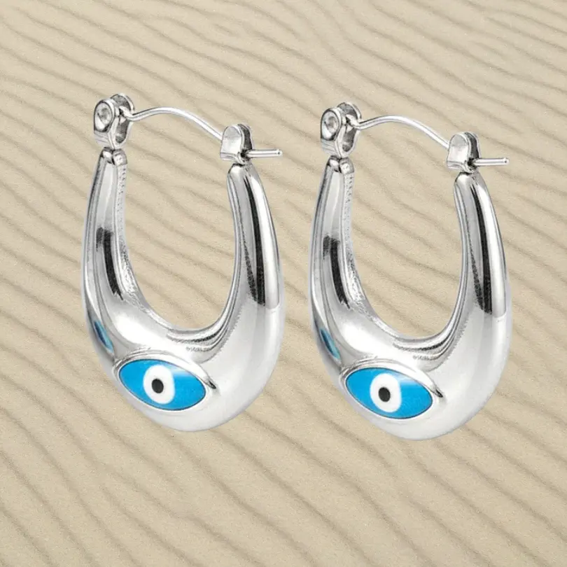 Boucles d'oreilles Oeil Oriental 1 Boucles d'oreilles Oeil Oriental en acier inoxydable finition mate argentée, motif œil protecteur, posées sur fond de sable