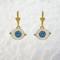 Accueil 12 Boucles d'oreilles pendantes œil protecteur doré, motif ovale bleu et blanc, posées sur fond de sable